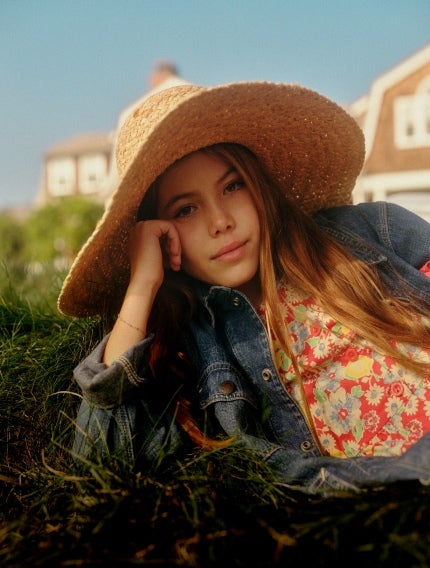 Girl wears a floral shirt, denim jacket, and straw sun hat.